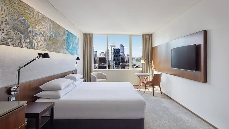 A photo of a room at Hyatt Regency, containing a queen bed with white sheets and a reading lamp, a marbled art piece, and a window with the cities buildings in the distance