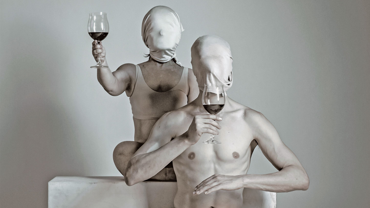 Two people painted white and wearing white hoods covering their faces hold up a glass of red wine each