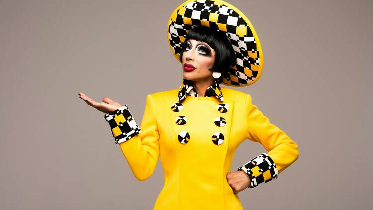 Bianca Del Rio holds her hand out to the side, as she poses for a photo in a yellow suit and hat