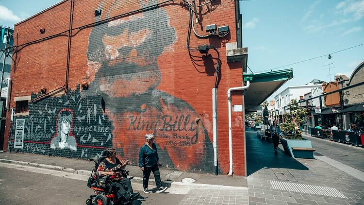 Two people (one in a wheelchair) stop in front of a large-scale mural in a laneway depicting an Indigenous man.