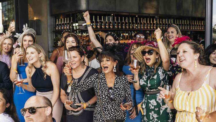 A group of excited people wearing fancy clothes and fascinators standing together and cheering.