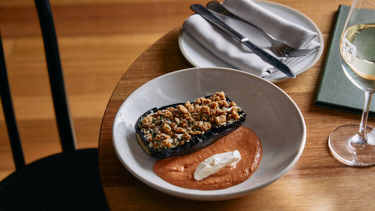 A spiced eggplant dish with mole sauce in a white bowl, atop a dining table.