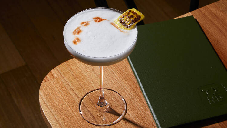 A Pisco Sour cocktail with a garnish branded with the word 'Tino'.