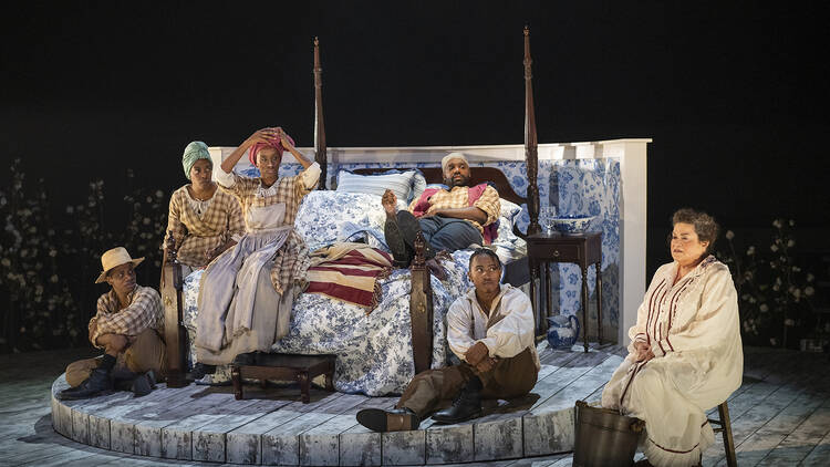 Actors portraying enslaved people sit on a bed looking at an actress, sitting in a chair, who's portraying Martha Washington