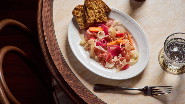 Prosciutto and colourful pickles sit on a white plate with toasted bread on a marble table