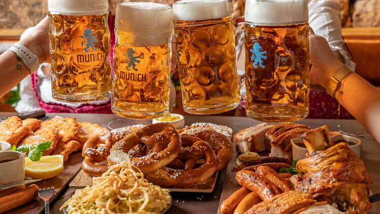 Four steins of beer held above a dining table topped with German treats like pretzels and sausages.