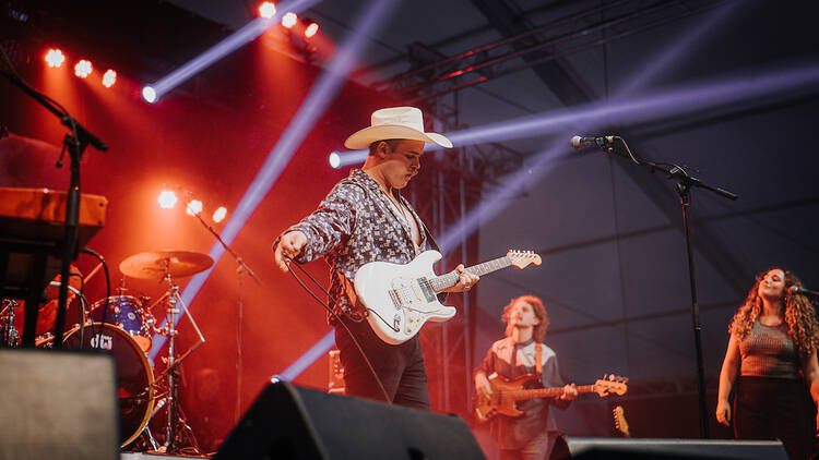 A man in a cowboy hat strums a guitar on stage with a band in the background. 