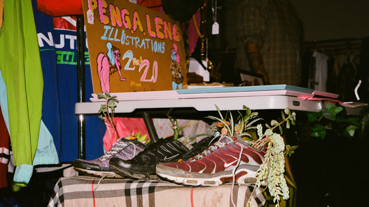 A few pairs of vintage shoes on a tartan rug, with streetwear on a rack in the background.