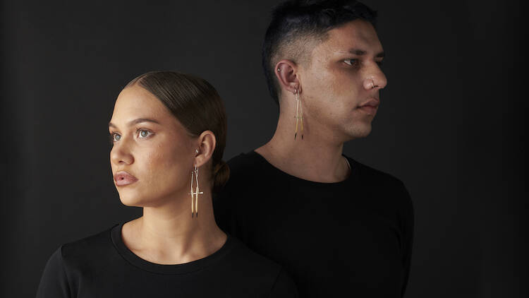 A woman stands in front of a man in front of a black background, both wearing black clothing and long gold jewellery front their ears.