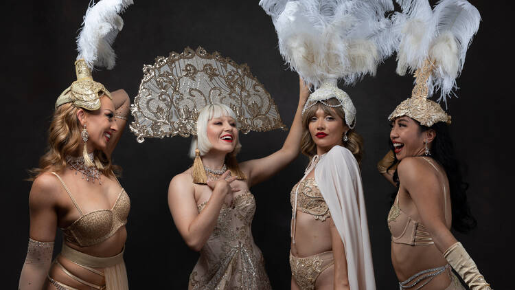 Four women dressed in white and gold burlesque costumes smiling and posing.