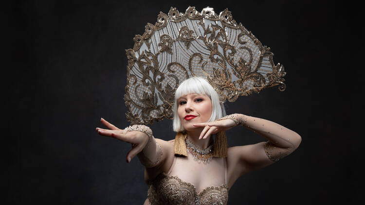 A women in a white wig dressed in a gold burlesque costume posing towards the camera.