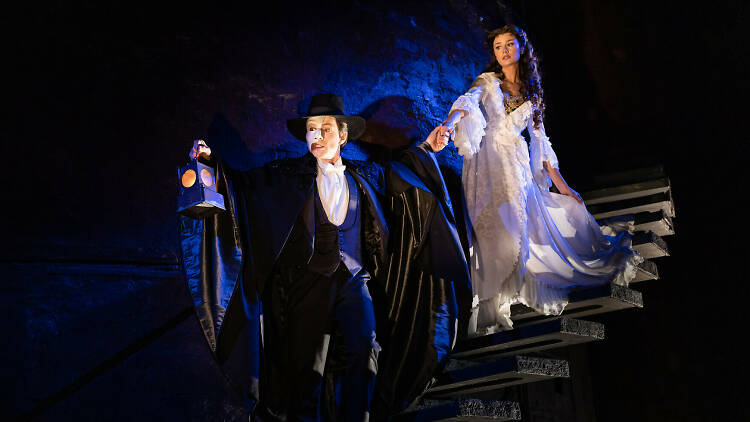A man in a mask leads a woman down stairs into the dark