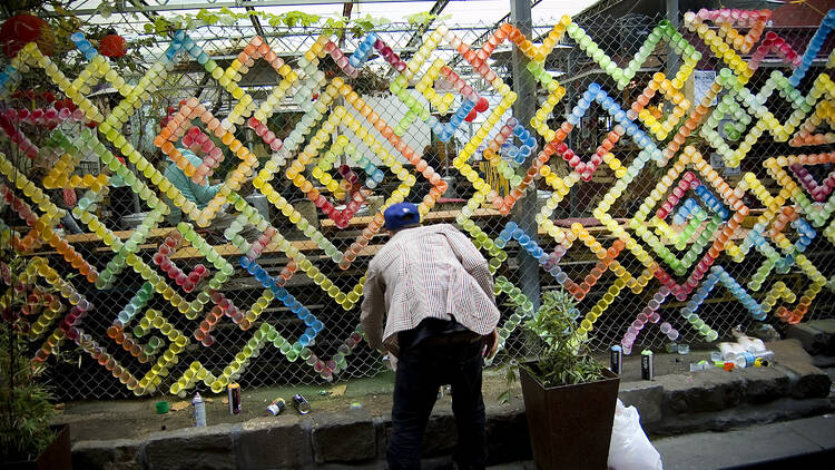A man in a white blazer stands in front of a street art style installation with colourful cups interlinked on a wire fence.