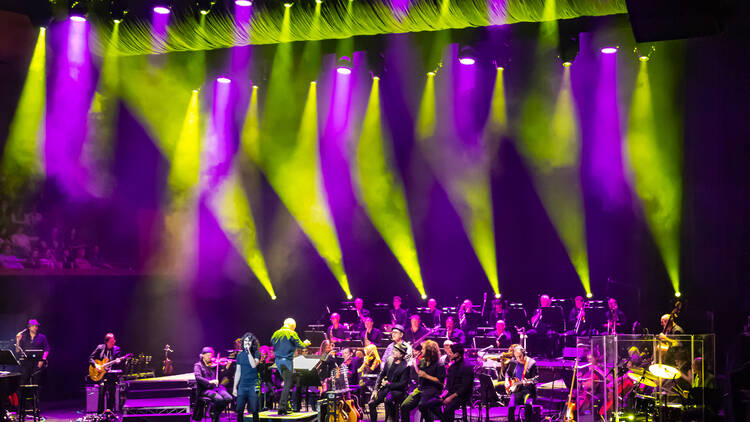 An orchestra and singers performing on stage surrounded by green and purple lighting.