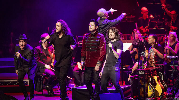 Four men singing on stage with an orchestra performing behind them bathed in red and purple lighting.