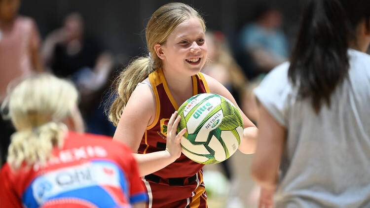 A smiling blonde girl gets ready to pass a netball on the court.