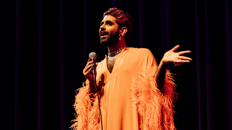 Alok Vaid-Menon holds a microphone on stage and wears an orange gown with a feather trim
