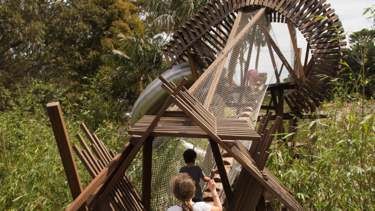 Kids run up a wooden boardwalk towards a crazy treehouse in the middle of a field