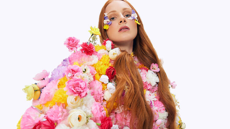 A woman with long orange hair looks into the camera and is covered by pink, white and yellow flowers.