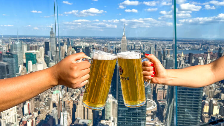 Two beer steins clinking with the NYC skyline in the background.