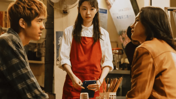 A man and a women sit look across a restaurant table at a each other while a waitress in a red apron stands by.