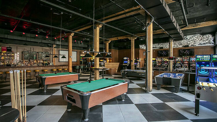 Arcade games and pool tables sit in a warehouse-like bar space with exposed ceilings and black-and-white checkered flooring