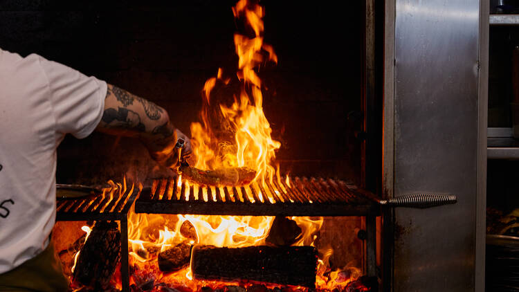 A chef cooking a slab of meat atop a woodfire grill.