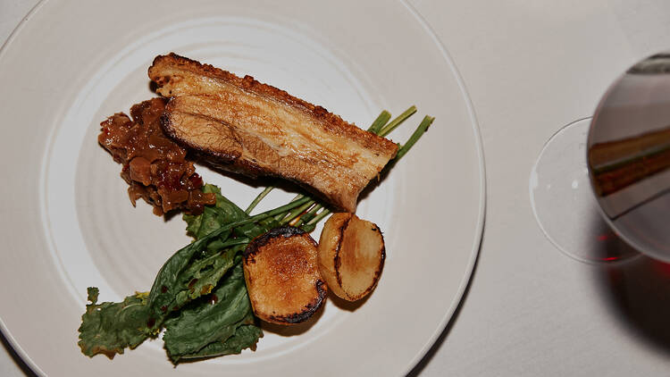 A slice of pork belly alongside greens and roasted potatoes.