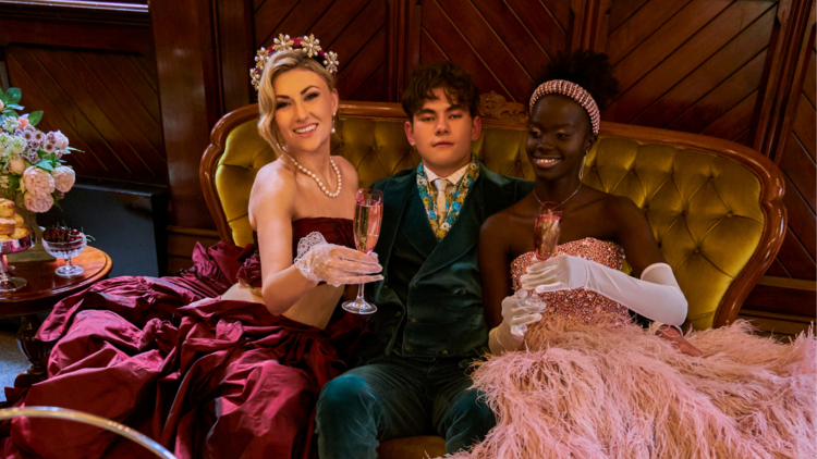 Three people sit on a velvet couch in ball gowns drinking champagne