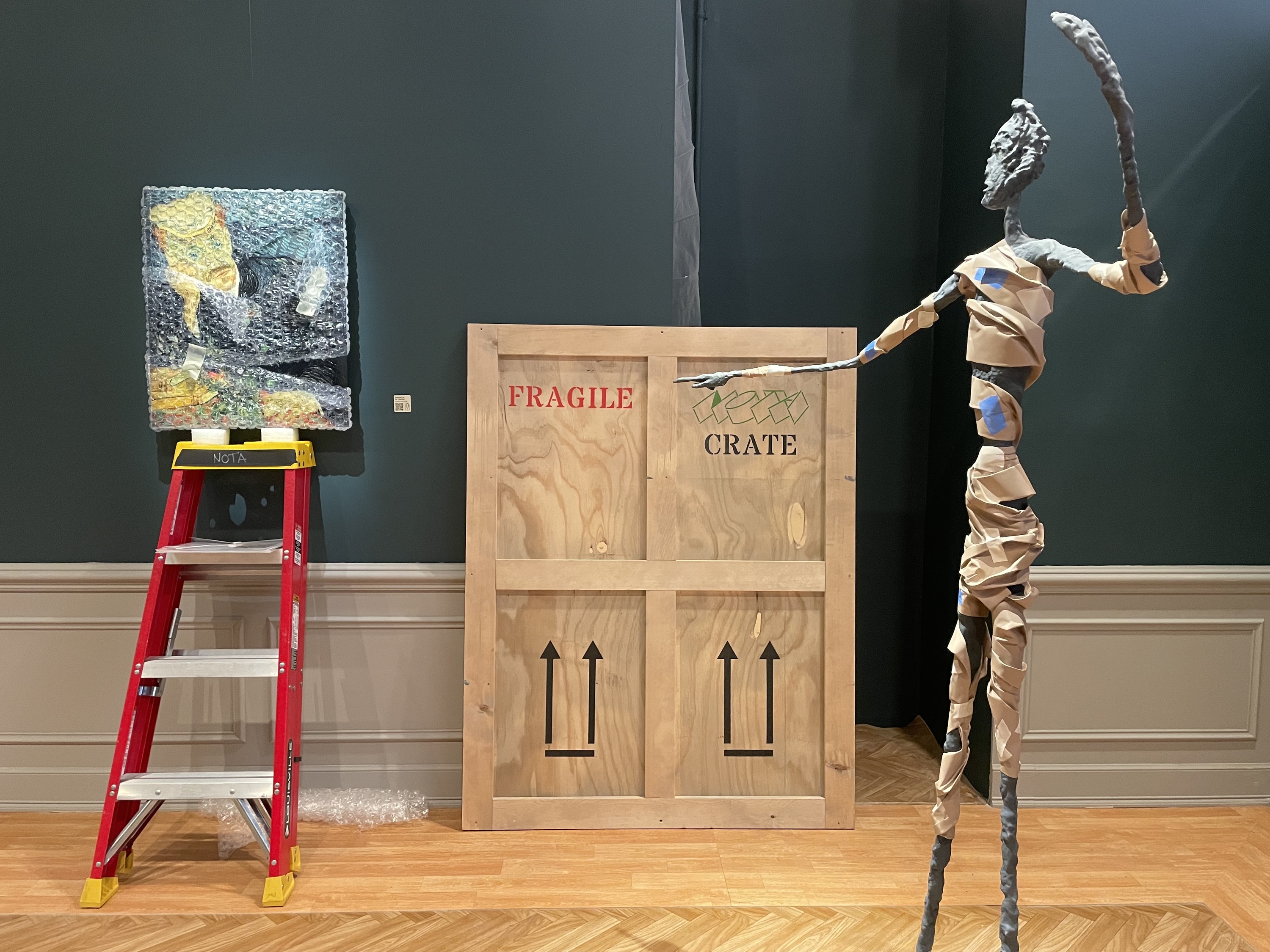 A painting sits on a ladder and a sculpture stands in front of a wooden art crate.