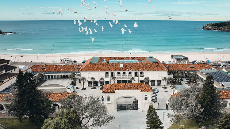 Aerial shot of Bondi Pavilion with birds flying over it along Bondi beach