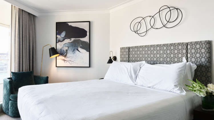 Two girls sit on a bed in a hotel room with water views eating room service 