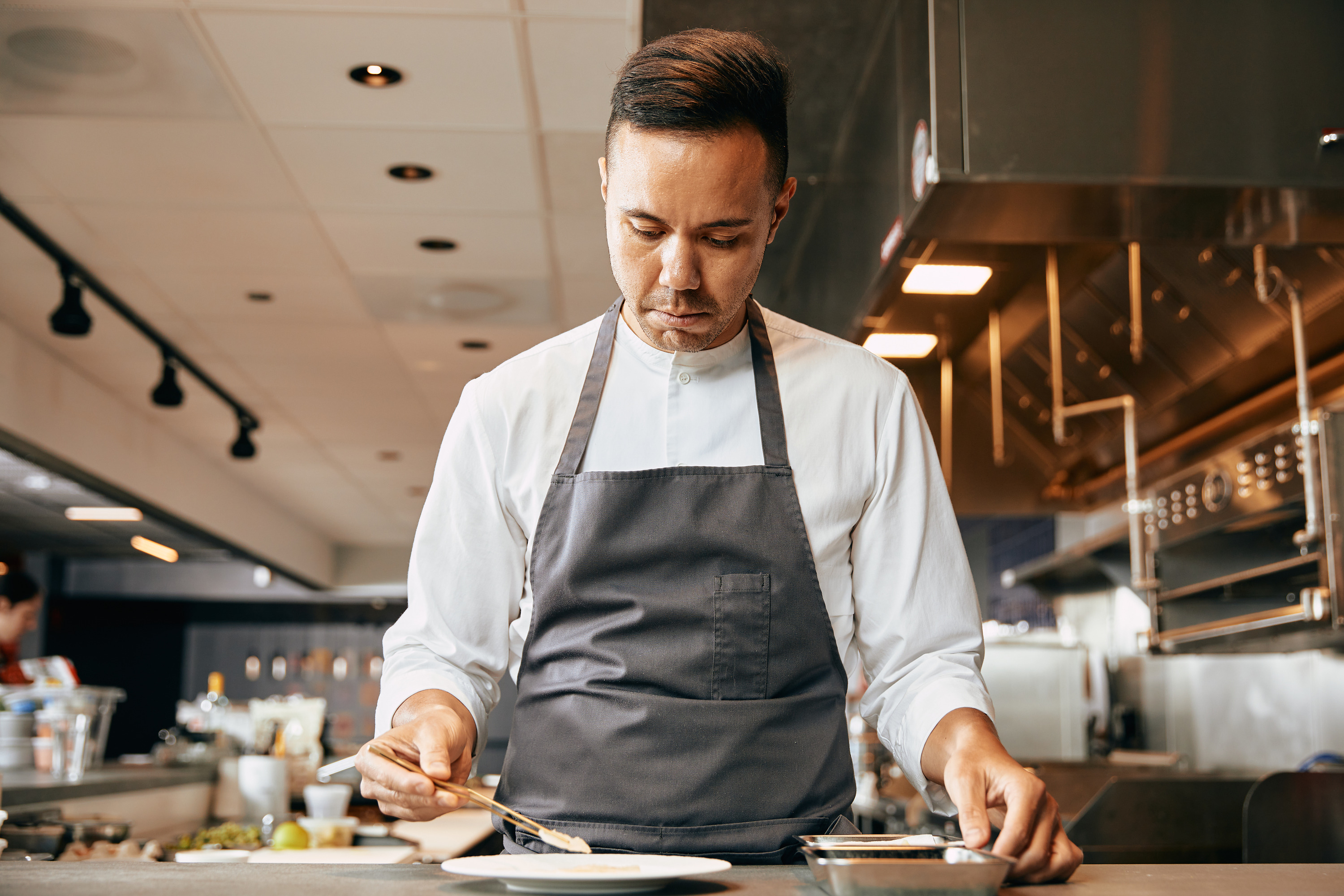 A man in a gray apron stands at a counter and plates a dish using chopsticks