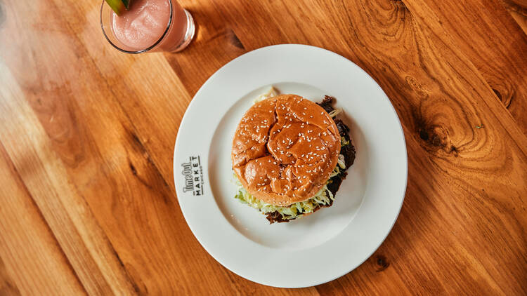 A smash burger with lettuce and a sesame seed bun sits atop a white plate on a wooded table next to a pink slushie drink