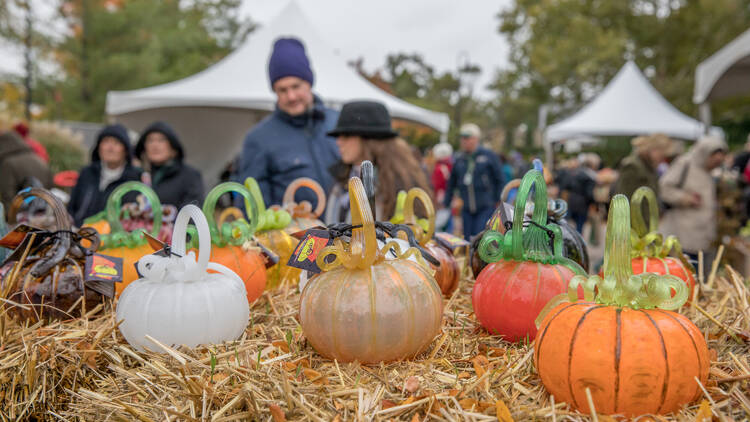 White, gold and orange pumpkins made of blown glass sit on top of a hay bale while people browse in the background