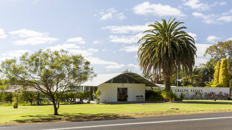 The front of Gralyn Estate, picturing a white building with a large palm tree next to it, bright green grass, and a visible road.