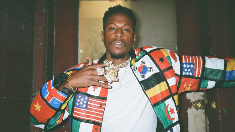 Joey Badass wearing a chunky gold necklace and leather jacket with flags on it.