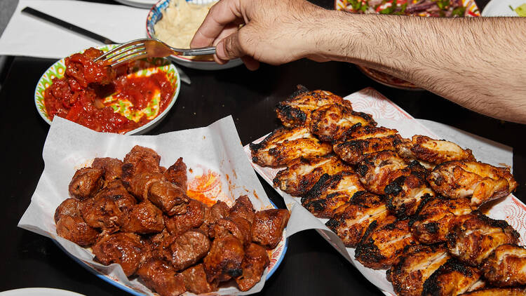 Someone reaches for a bowl of food, with a selection of dishes on a black table including chicken and kebabs