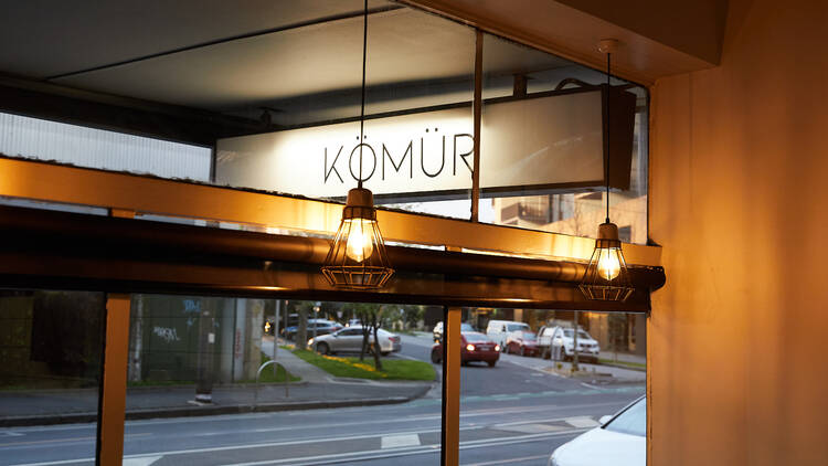 A white sign outside the restaurant reads Komur with hanging lights inside giving a golden colour