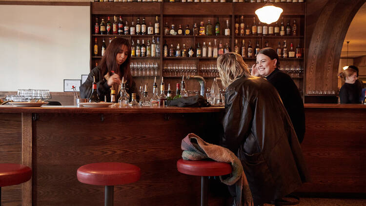 Two friends sit at a wooden bar on red leather stools while a bartender makes a cocktail behind them