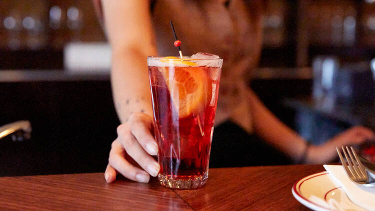 A bartender places a red coloured cocktail with an orange slice inside on a wooden bar