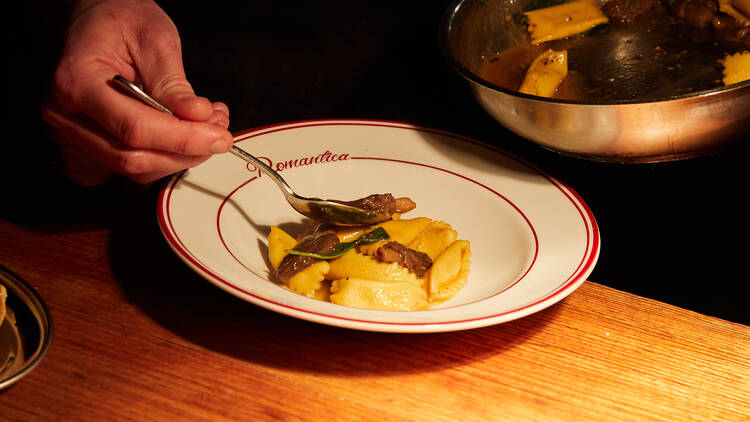 A chef spoons pasta into a white bowl on a wooden bench