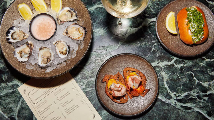 A plate of oysters on ice and other small snacks sit with a glass of white wine on a green marble bench