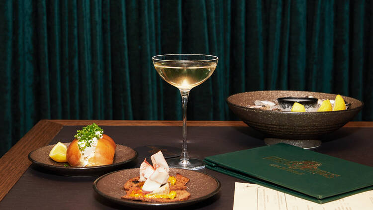 On a table in front of a dark green velvet curtain there is a cocktail glass, a menu, and three plates of food