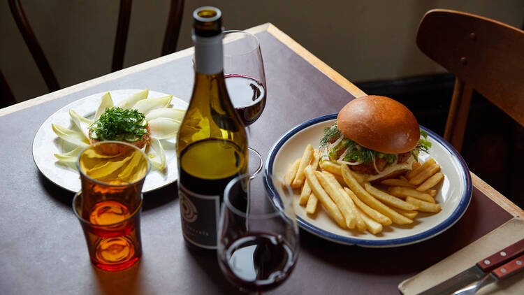 A dark table with a bottle of red wine and two red wine glasses, a fish burger and chips and trout rillette dish with two orange water glasses