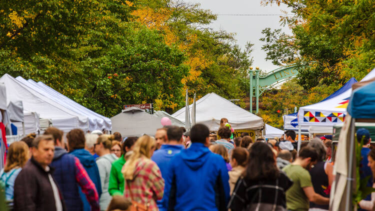 A crowd of people walking down a street lined with white tents and leaves with green and yellow leaves