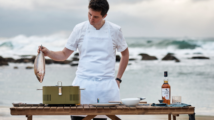 Chef Josh Niland holding up a fish over a grill on the beach