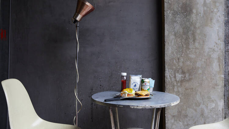 Two white chairs sit next to a small table topped with a burger, ketchup and napkins, next to a light fitting against a dark grey wall