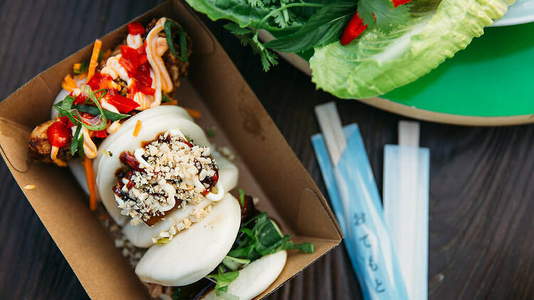 Filled bao buns sit inside a cardboard box with chopsticks on the table next to them 