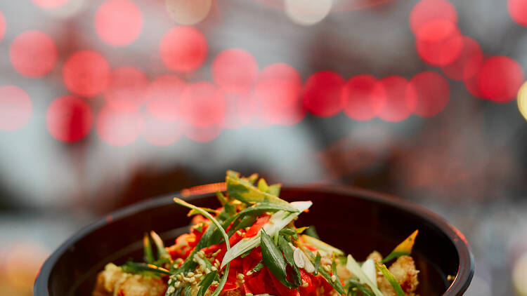A bowl of noodles topped with spring onions and chilli sits in front of blurred red lights